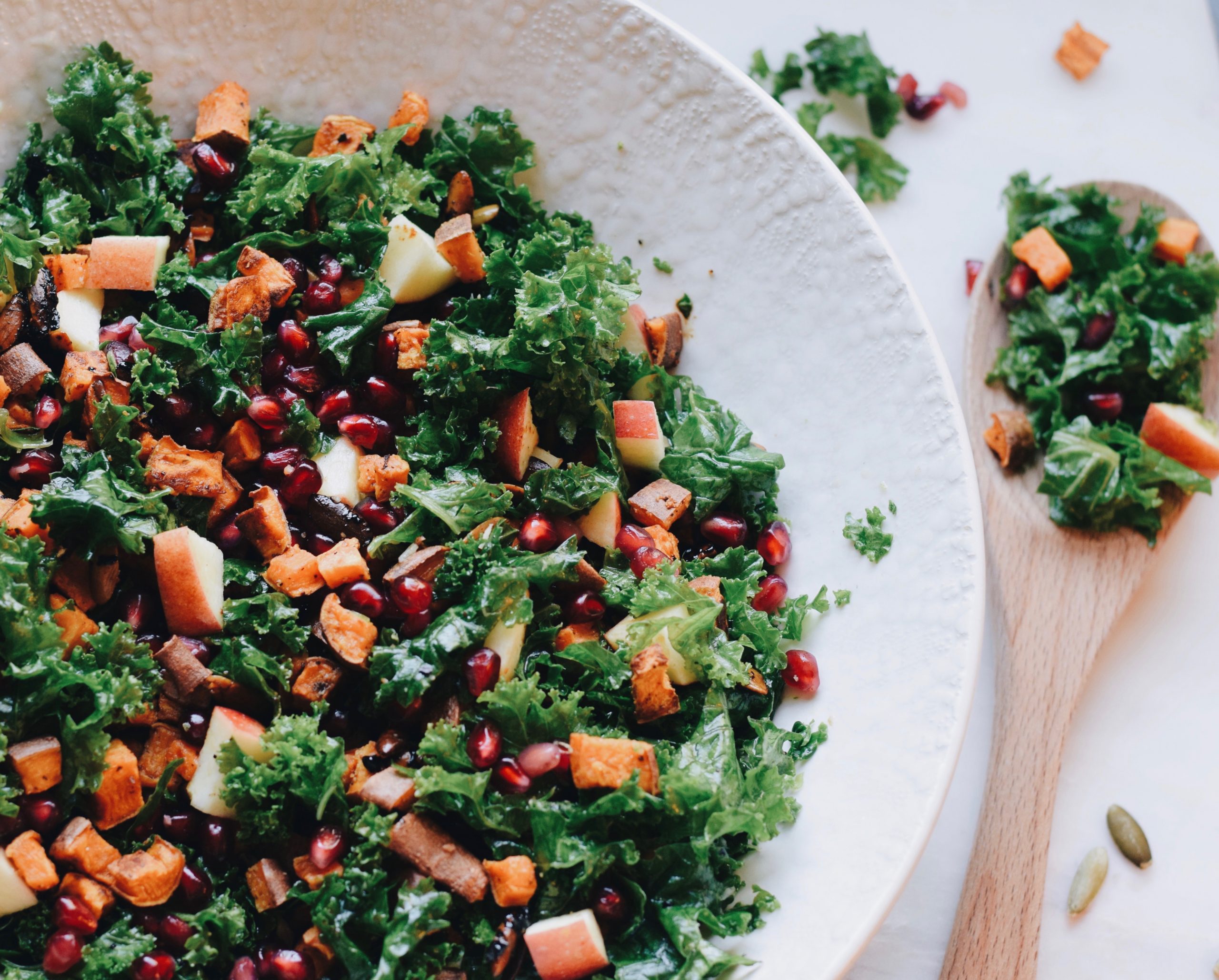 kale salad with apples, sweet potatoes, pumpkin seeds and pomegranate seeds in a white serving dish